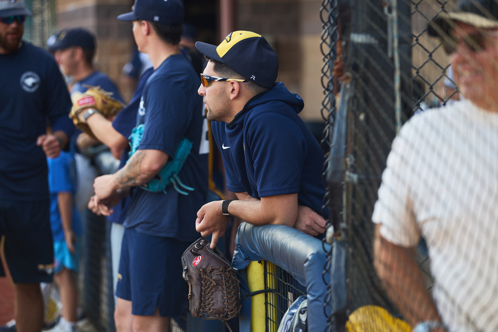 A baseball player in a navy hoodie and a gold-brimmed cap leans on the dugout railing, focused on the field. He holds a brown leather glove in his left hand.