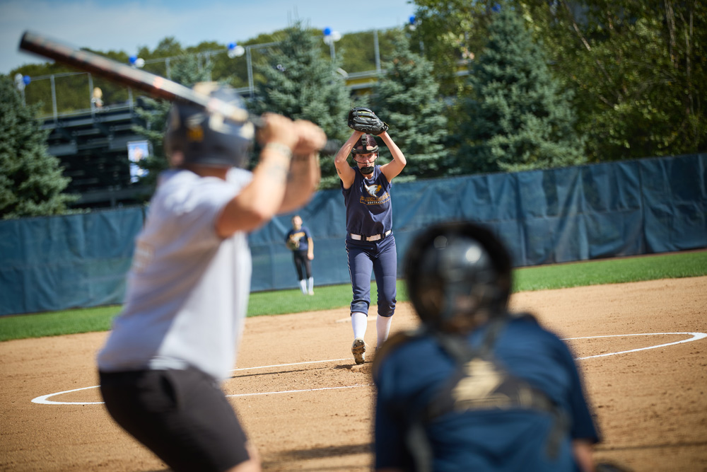 A view from behind the catcher shows a softball pitcher in mid-delivery on a dirt mound. A batter stands ready at the plate against a background of evergreen trees.