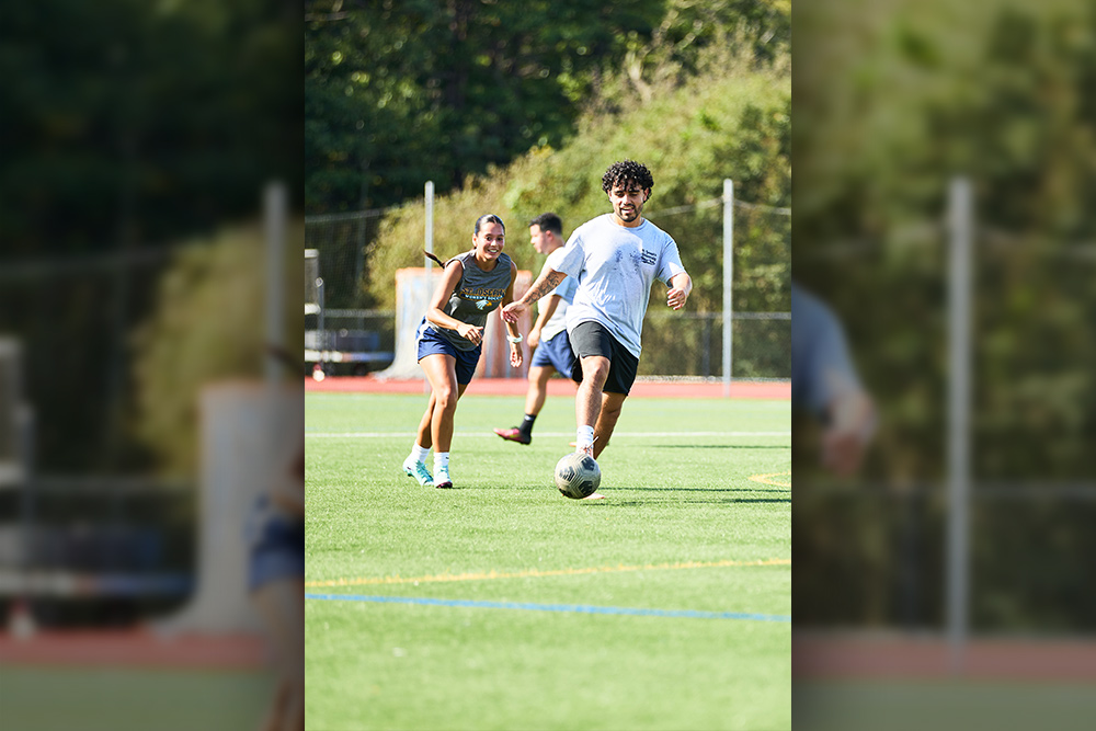 A young man with curly hair dribbles a soccer ball down a green turf field during a match, while a female player in a gray "St. Joseph's Soccer" shirt follows close behind.