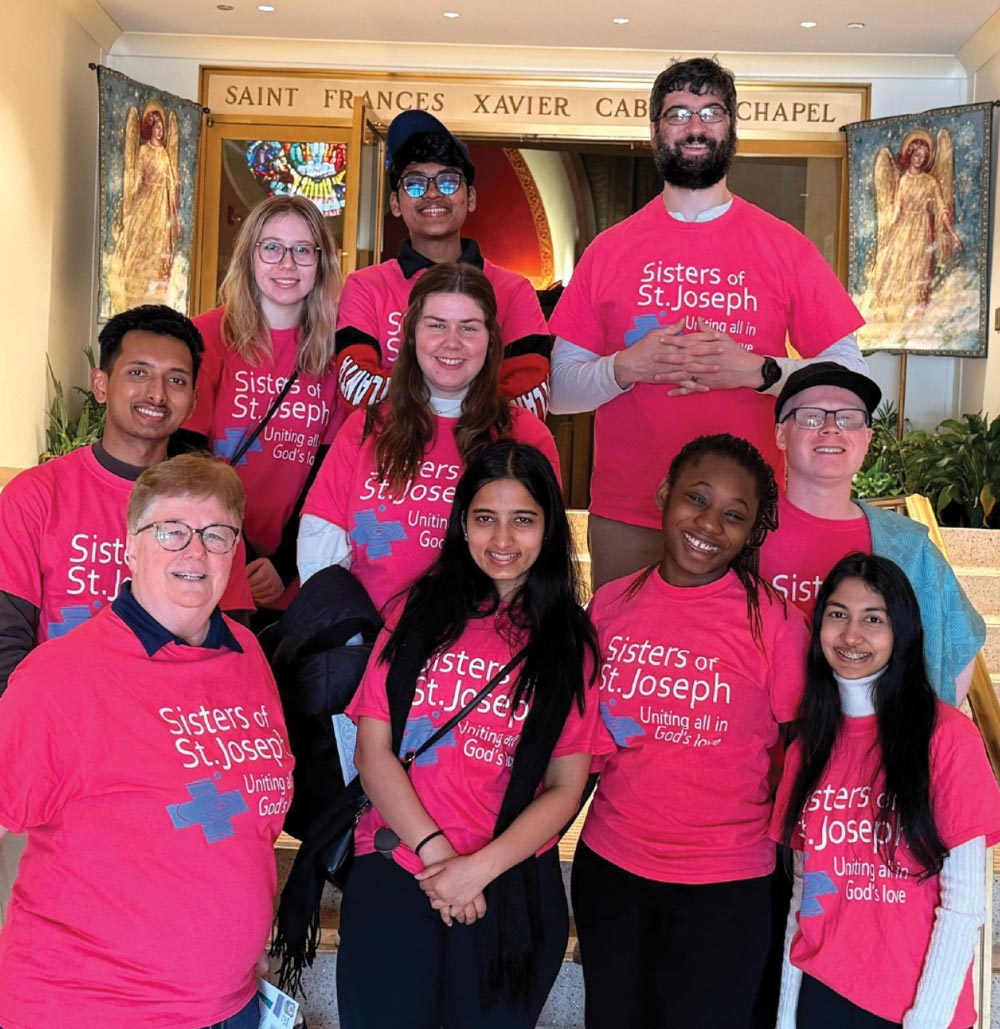 A group of diverse students and a leader pose on stairs in front of a chapel. They are all wearing matching bright pink t-shirts that read "Sisters of St. Joseph: Uniting all in God’s love."