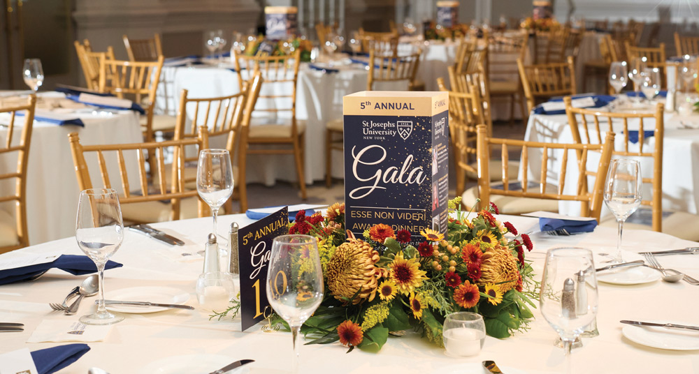 An elegant banquet hall setup with round tables, white linens, and gold chairs. Each table features a floral centerpiece and a navy blue "5th Annual Gala" program for St. Joseph’s University.