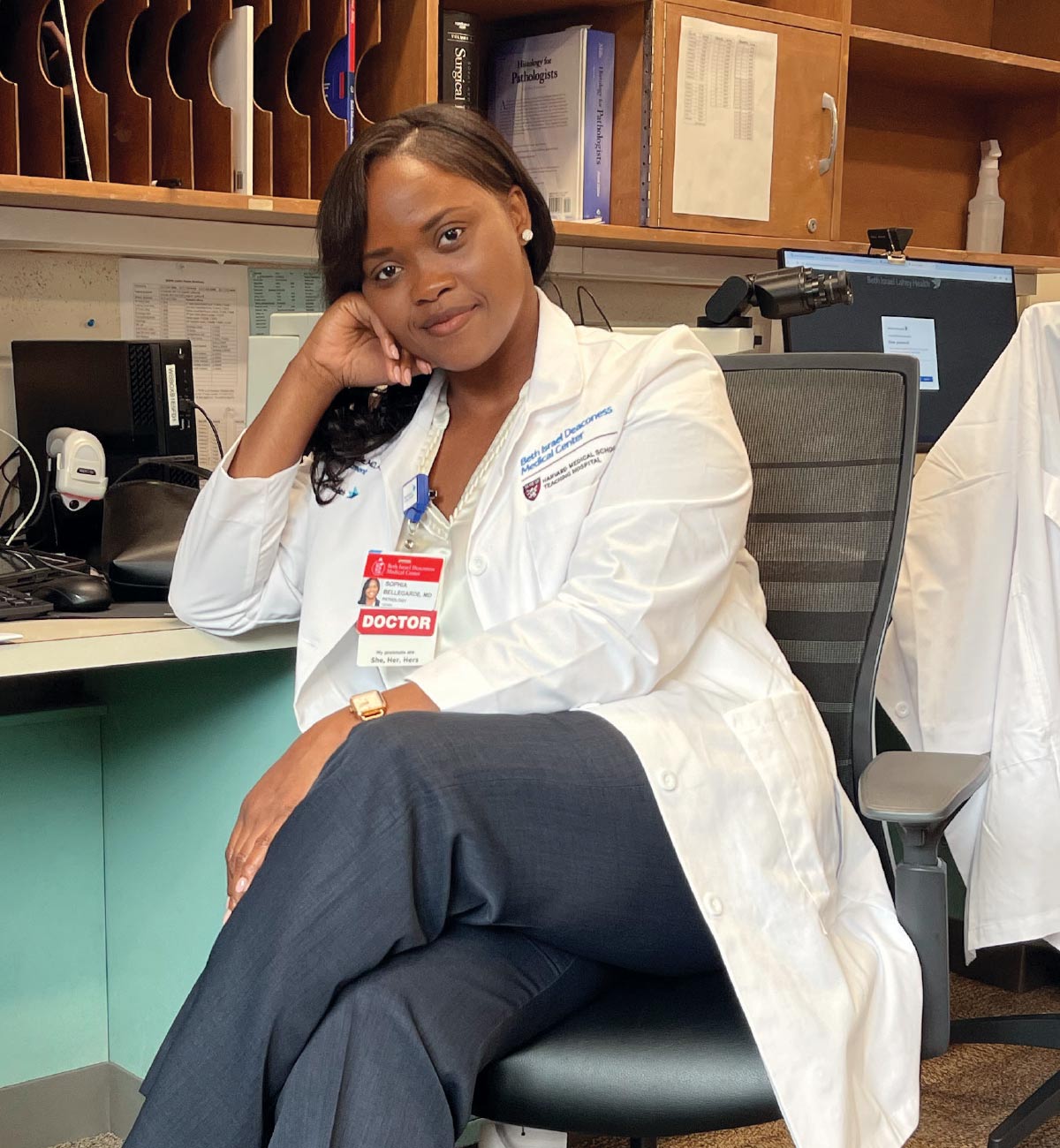 Sophia Bellegarde in a white doctor's coat sitting at a desk in a medical office, leaning her head on her hand.