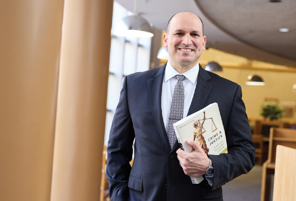 Jason Ostrowe in a pinstripe suit and tie standing in a library while holding a book titled "Crime & Justice."