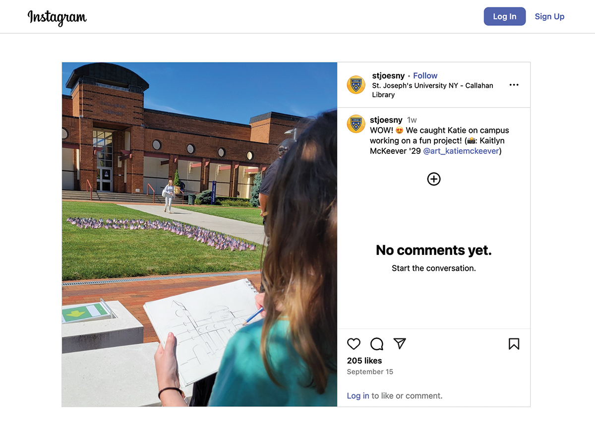 A screenshot of a Instagram post from the account labeled stjoesny showing a student on campus in front of the Callahan Library; In the foreground, the student’s hands are shown sketching the library building in a notebook, while in the background, a lawn is decorated with hundreds of small American flags arranged in a pattern