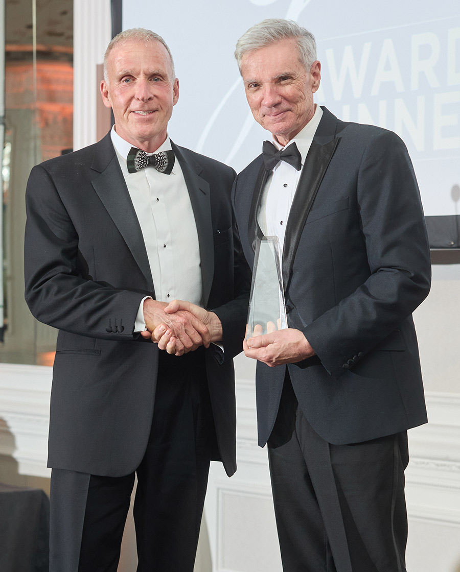 Two men in tuxedos shaking hands. The man on the right holds a clear, triangular glass award. They are standing indoors in front of a screen displaying the word “AWARD.”