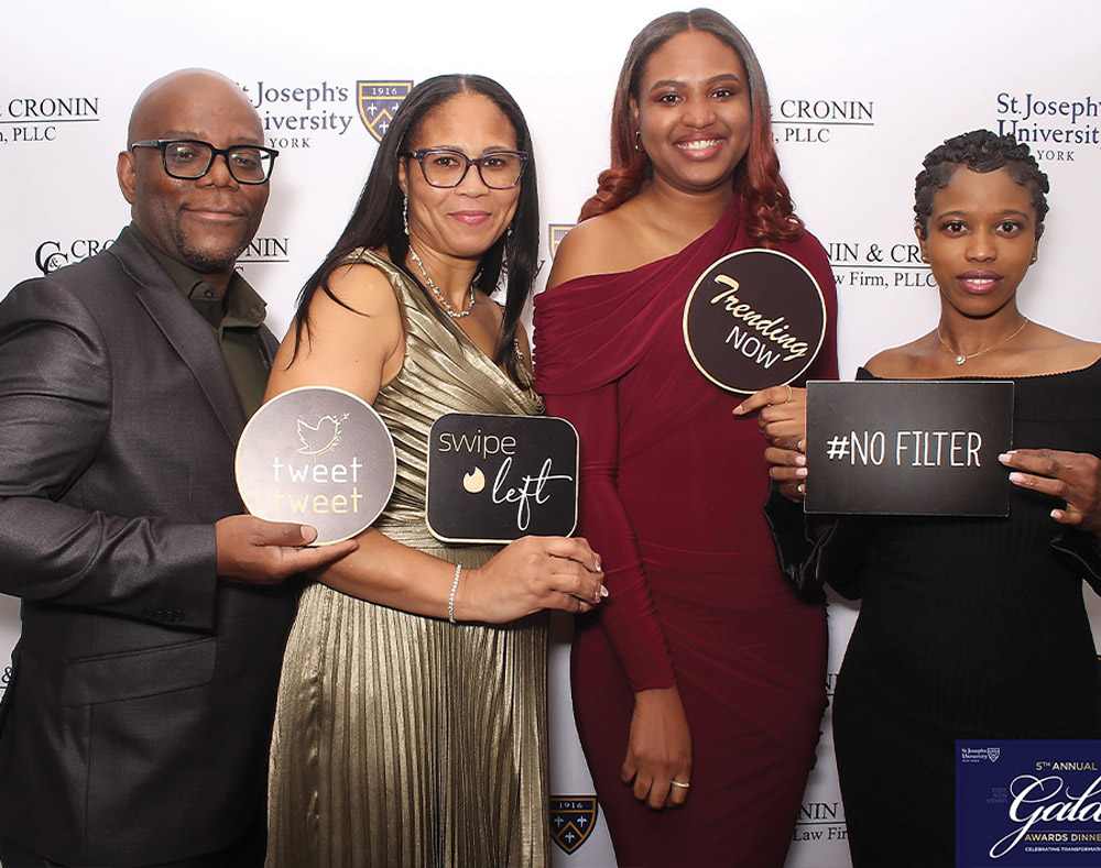 Four people pose in front of a university-branded backdrop while holding social media-themed signs that read “tweet tweet,” “swipe left,” “Trending NOW,” and “#NO FILTER.” They are dressed in formal evening wear and smiling.