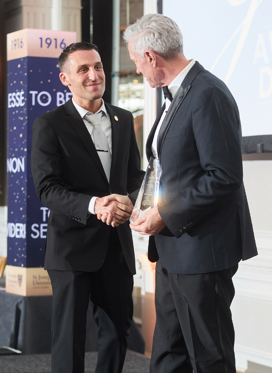 A man in a black suit and silver tie shakes hands with a man in a tuxedo holding a clear, pointed award. Behind them is a decorative pillar with the university motto "Esse Non Videri."
