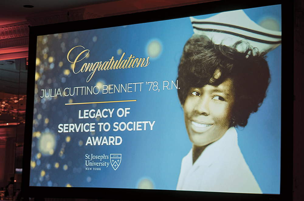 A projection screen displays a vintage photo of a woman in a nurse’s cap. The text includes “Congratulations” and “Legacy of Service to Society Award” alongside the St. Joseph's University logo on a decorative blue background.