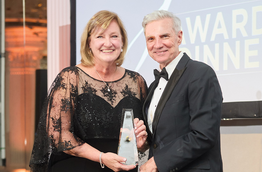 A woman in a black formal gown with lace sleeves and a man in a tuxedo smile while holding a clear, triangular glass award together. They stand in front of a projection screen displaying the words “AWARD DINNER.”