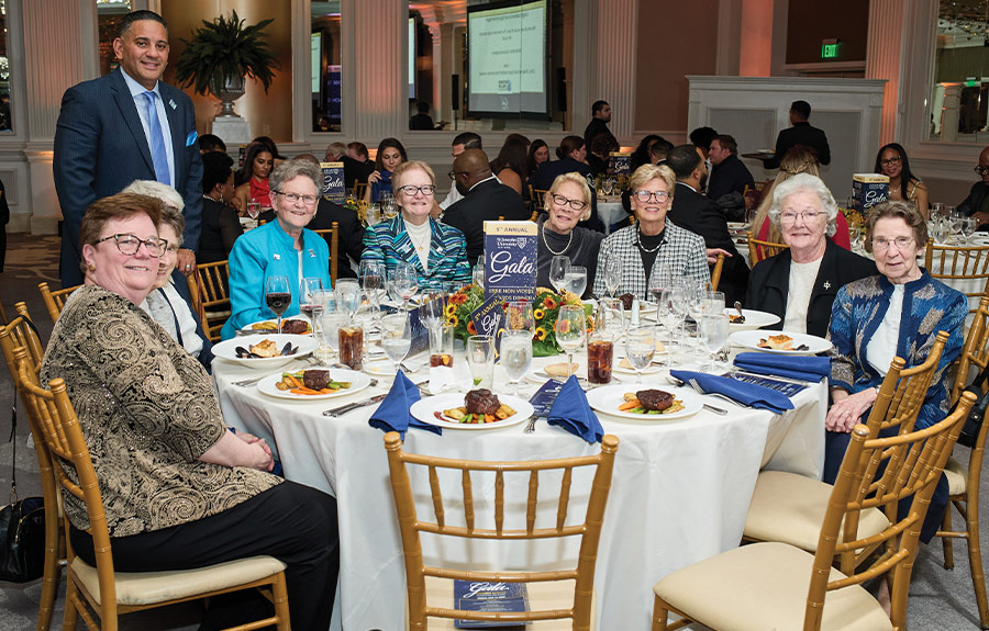 A group of women and one standing man smile around a gala dinner table set with white linens and steak entrees. A blue event program stands as a centerpiece in a crowded ballroom with a projection screen in the background.