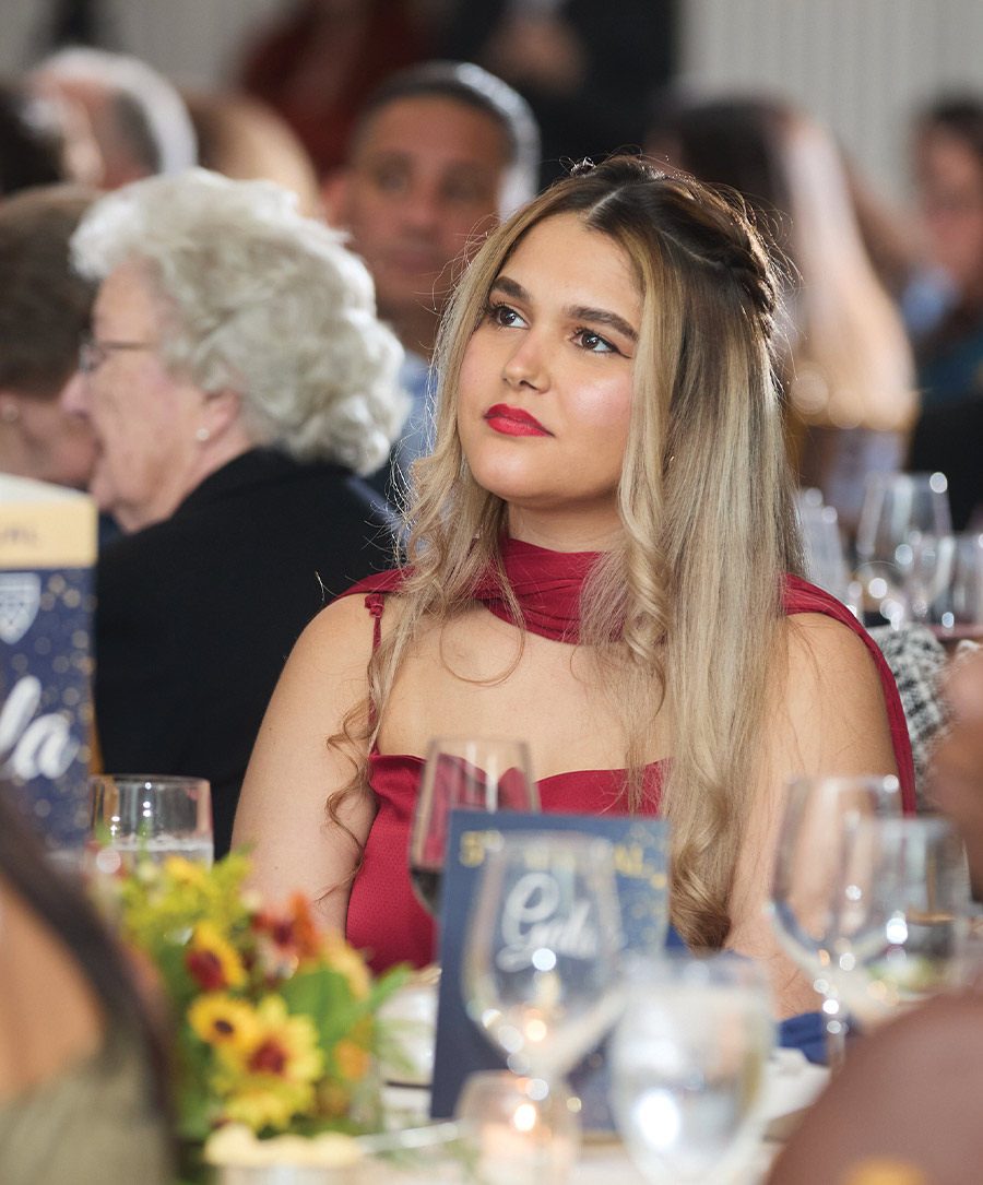 A woman in a red formal dress and red lipstick sits at a gala table, looking upward with a slight smile. The table is set with floral centerpieces and blue event programs, with other guests blurred in the ballroom background.