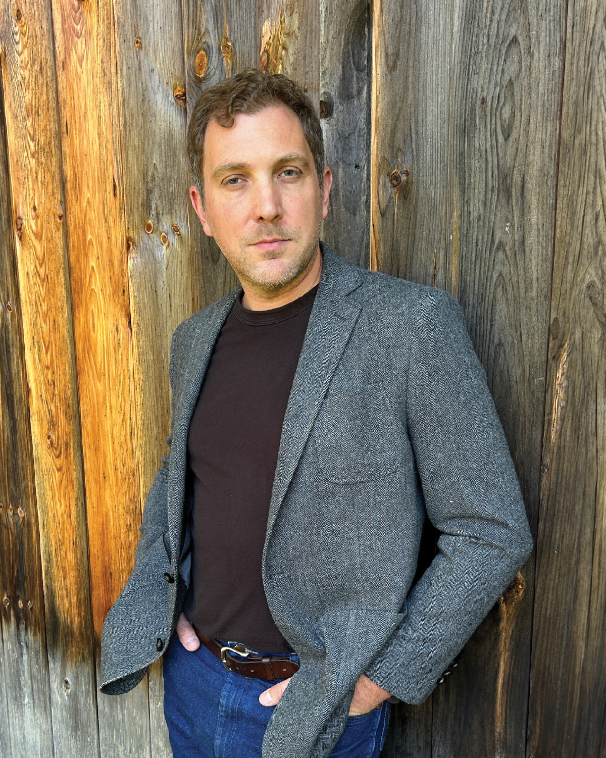 A portrait of Lee Clay Johnson, a man with light brown hair and a slight beard, is seen wearing a brown shirt and a gray herringbone blazer suit; He is leaning against a weathered wooden wall and has his hands resting inside the pockets of his blue denim jeans