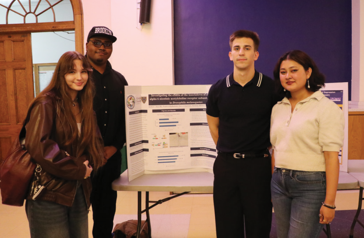 Four SJNY students stand together in a hall behind a white trifold poster board presenting research.